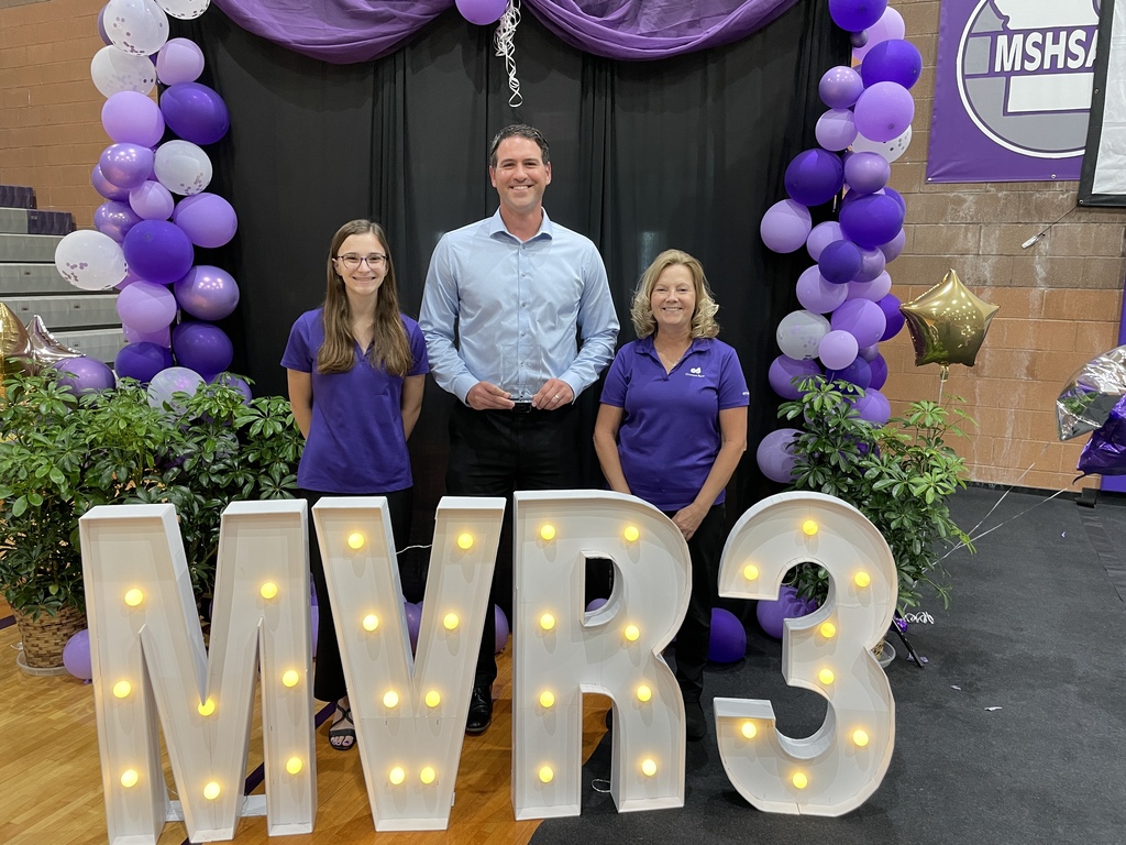 Three people pose behind illuminated “MVR-3” letters at an awards event, framed by purple balloon arches and decorative greenery.