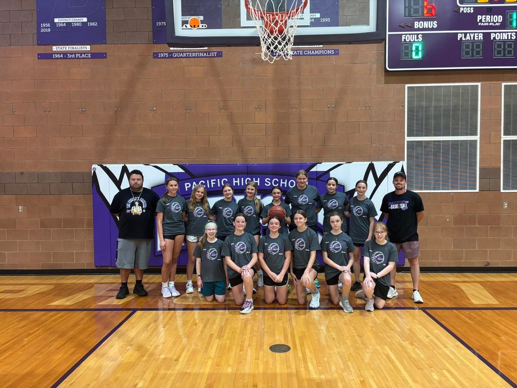 Girls basketball camp participants and coaches pose for a group photo on the Pacific High School gym floor beneath a basketball hoop.