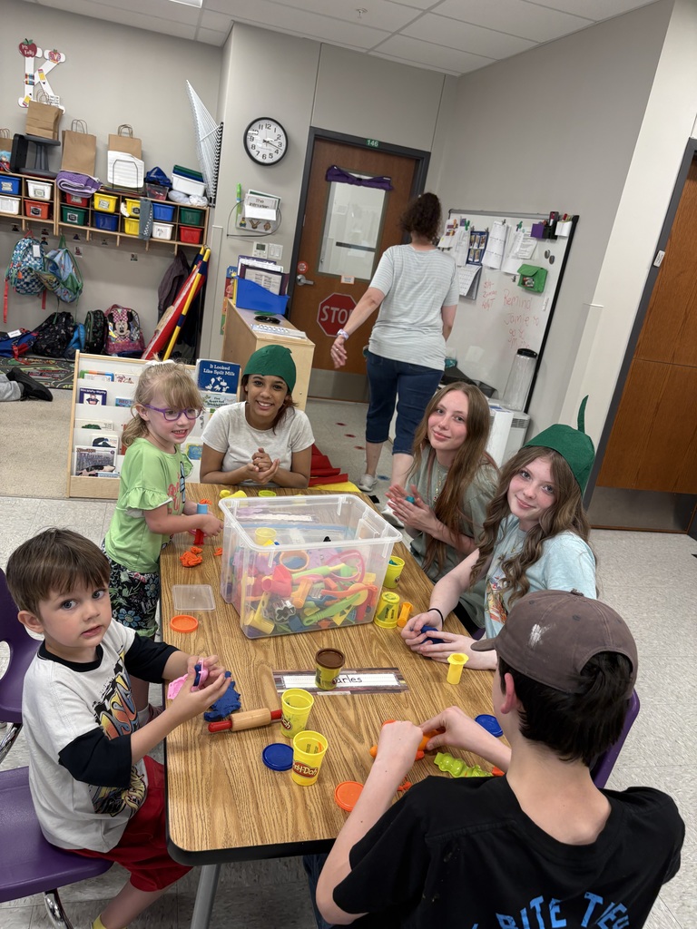 Children and teen helpers gather around a classroom table playing with colorful modeling clay and tools during a hands-on learning activity.