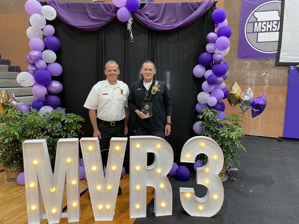 Two adults stand in front of a lighted "MVR3" sign.