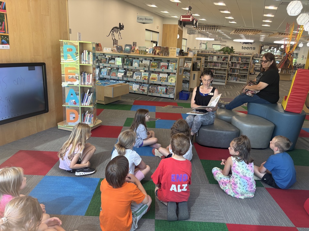 Children sit on a colorful library floor listening to a story during a read-aloud session, surrounded by bookshelves and reading displays.