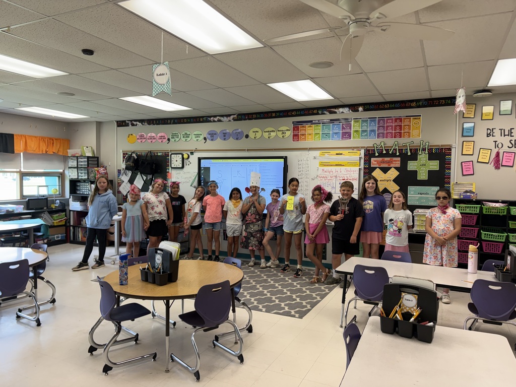 Elementary students line up across a classroom in colorful outfits and props, participating in a class activity while posing for a group photo.