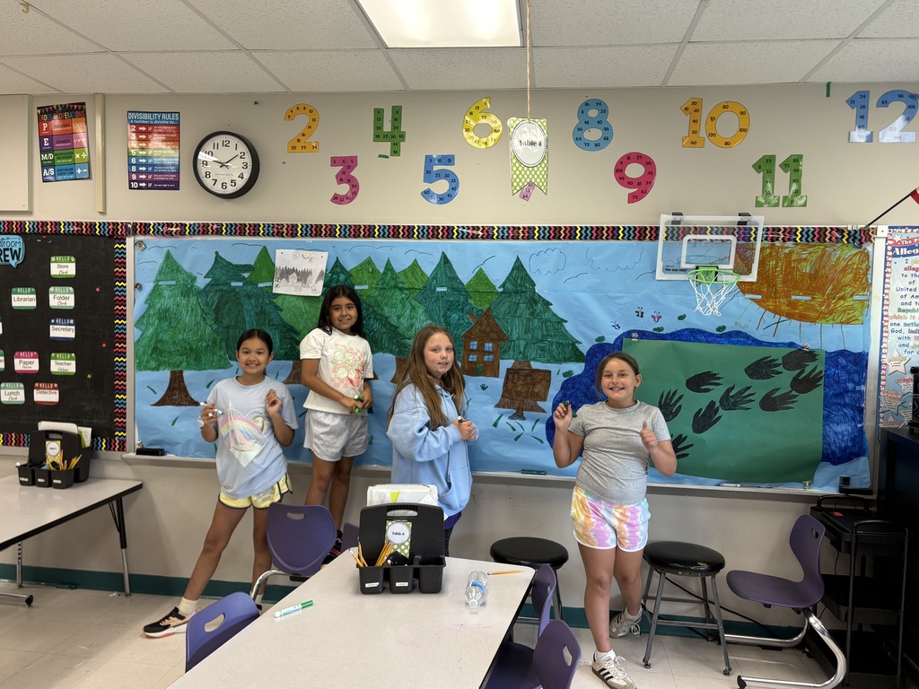 Four students stand in a classroom beside a large hand-drawn mural of a forest, cabin, and dinosaur tracks, proudly displaying their artwork.