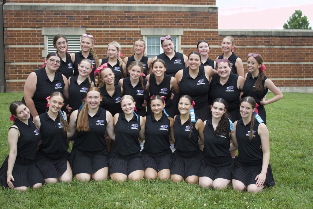 Pacific High School cheerleaders pose outdoors on a grassy lawn in matching black team outfits, smiling in front of a brick school building.