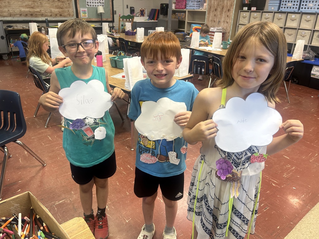 Three elementary students proudly display cloud-themed classroom projects with hanging drawings, standing together in an art-filled classroom.