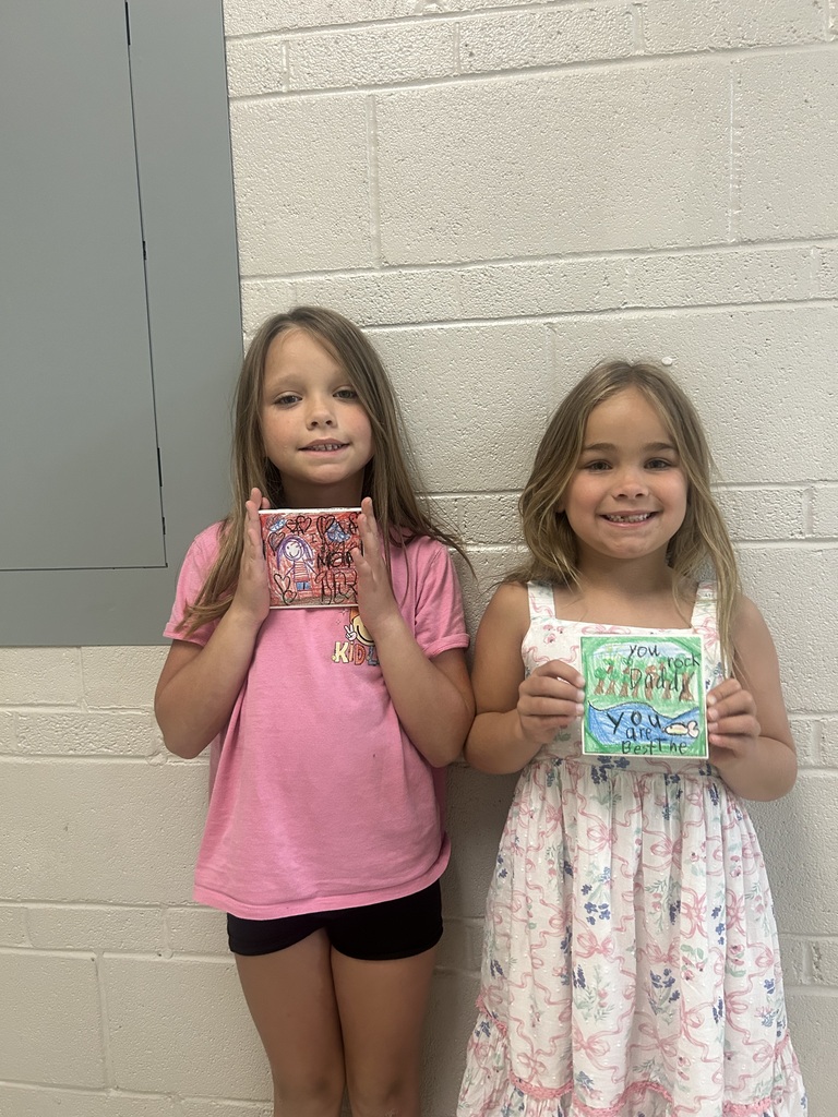 Two young students smile while holding colorful handmade artwork, standing against a white brick wall in a school hallway.