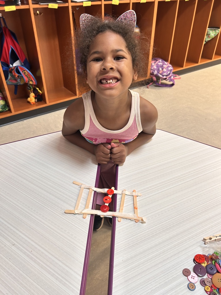 Young student smiles proudly beside a handmade bridge built from craft sticks and buttons during a classroom STEM activity.