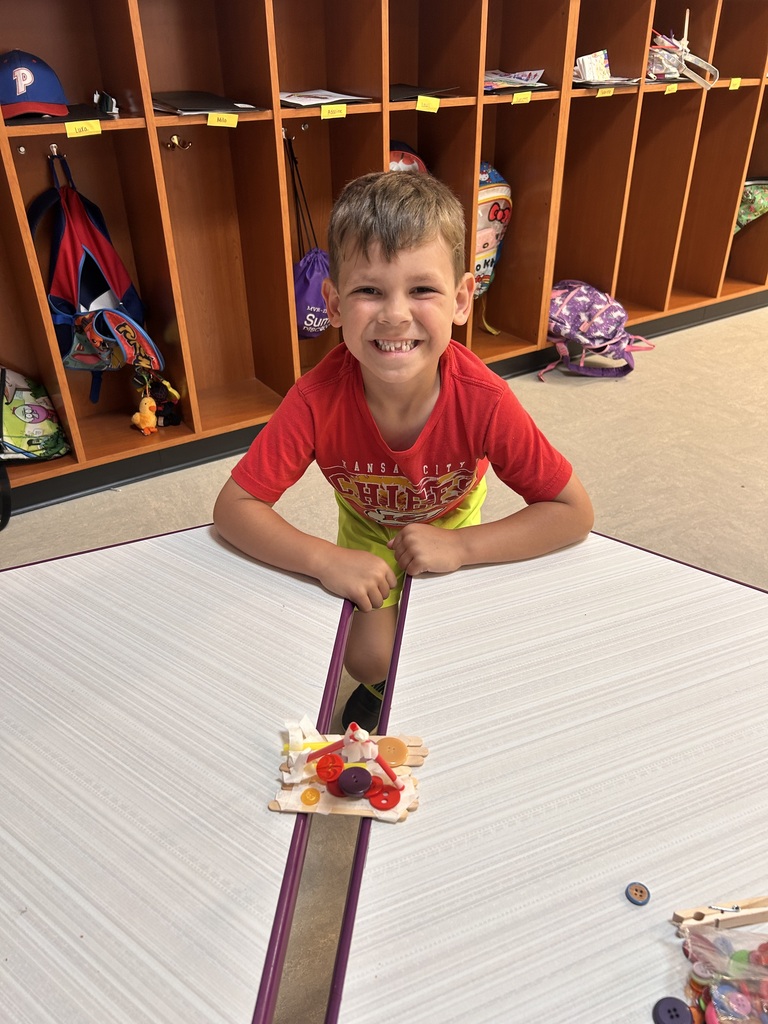 Student grins while presenting a craft-stick bridge decorated with colorful buttons, displayed between two classroom tables