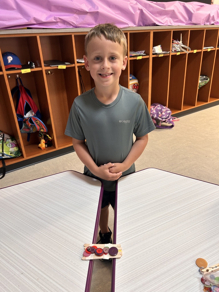 Student stands behind a completed craft-stick bridge, showcasing a classroom engineering project made with buttons and wooden sticks.