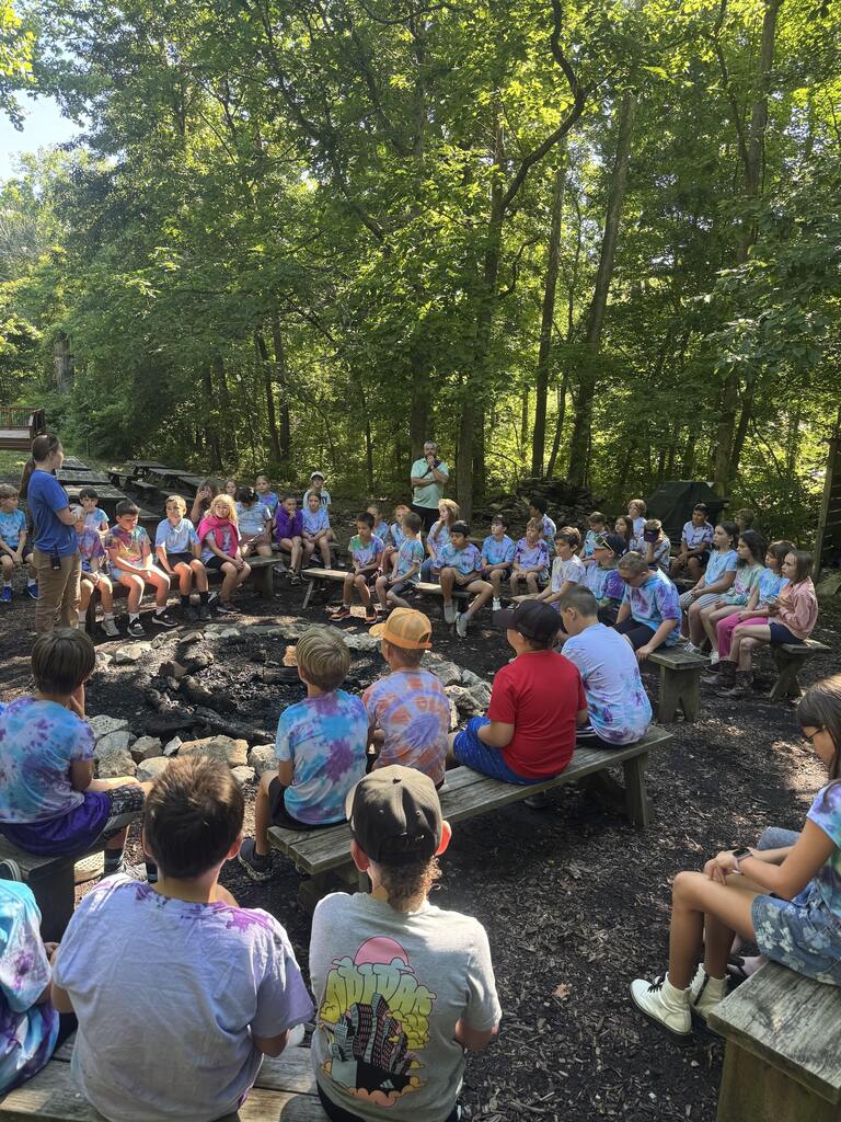 Students gather around an outdoor campfire circle, listening to a presenter during an educational nature program in a wooded area.