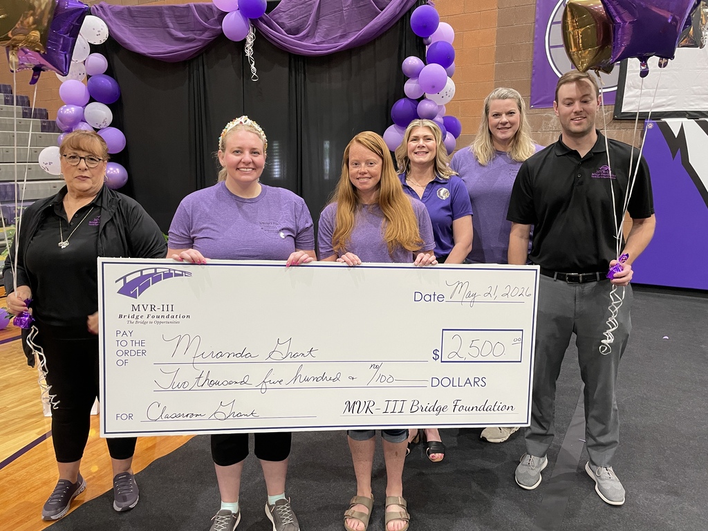 Six MVR-III staff members stand on a decorated stage holding a ceremonial $2,500 classroom grant check awarded to Miranda Grant by the MVR-III Bridge Foundation.