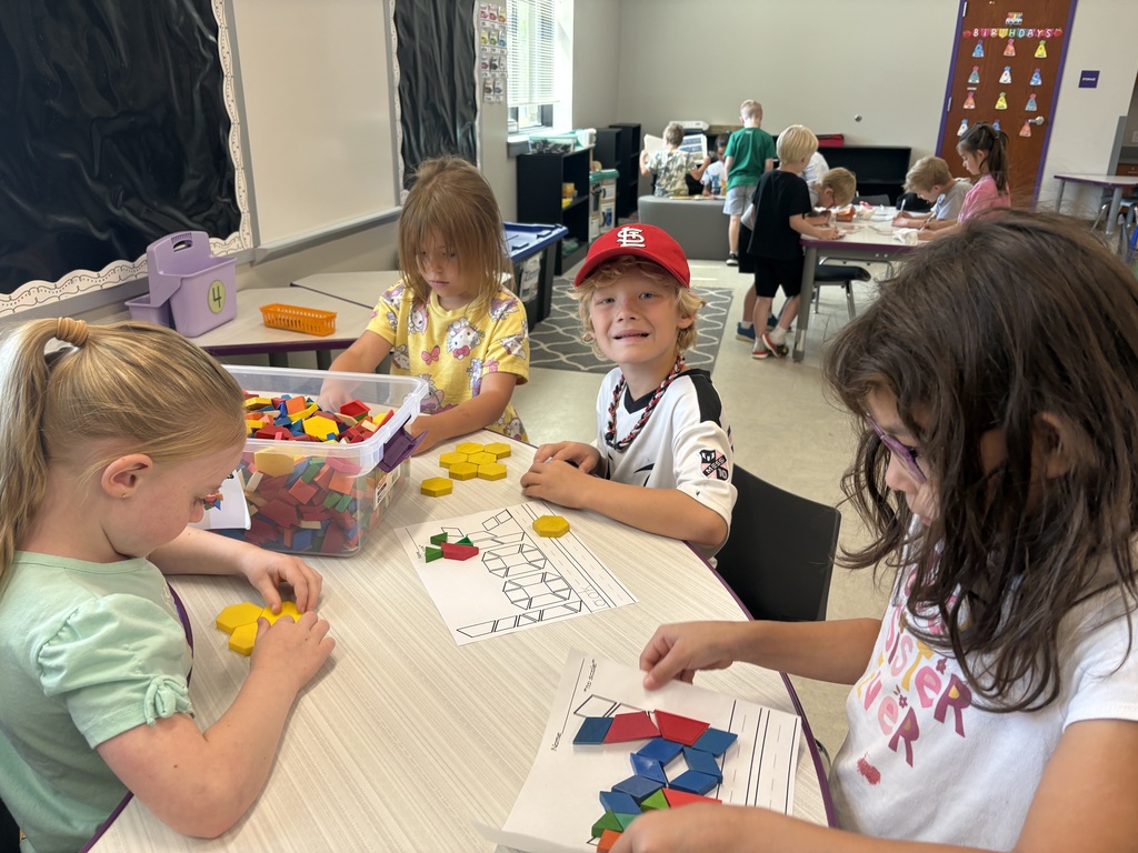 Children work with pattern blocks and shape activities at a classroom table while classmates engage in learning stations around the room.