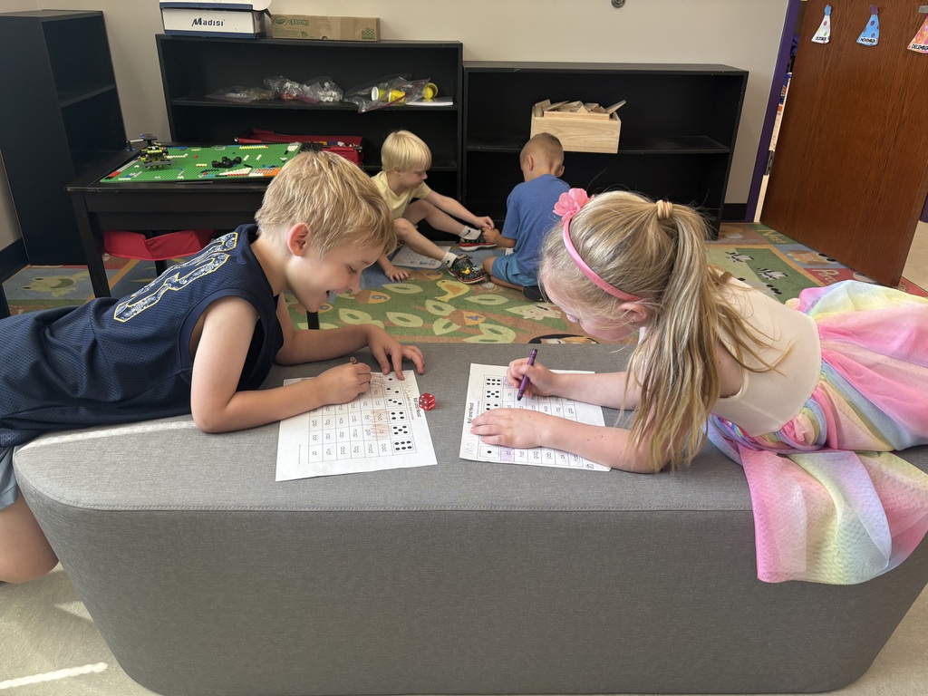Two young students play a math game with worksheets and dice on a cushioned bench while classmates build and explore nearby.