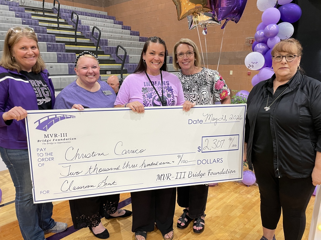 Five women stand in a school gym holding a ceremonial check from the MVR-III Bridge Foundation awarding Christina Carrico $2,307.91 for a Classroom Grant, with purple balloons in the background.