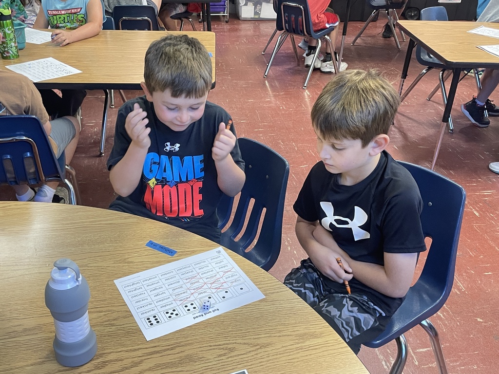 Students work together on literacy activities in a classroom, rolling dice, reading words, and completing worksheets at small-group tables.
