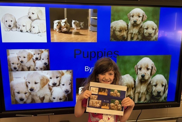 Student holds a printed project while standing in front of a classroom presentation about puppies, featuring multiple photos of young dogs on a large screen.