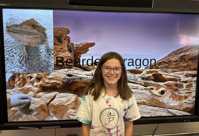 Student smiles in front of a classroom presentation about bearded dragons, with photos and illustrations of the reptile displayed on a large screen.