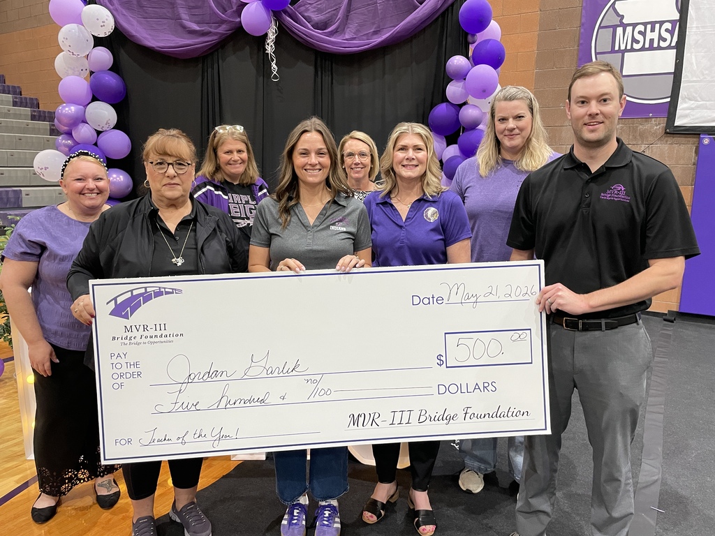 Group of educators and school leaders pose on a decorated stage with a large ceremonial check for $500 awarded to Jordan Harlik as Teacher of the Year by the MVR-III Bridge Foundation.