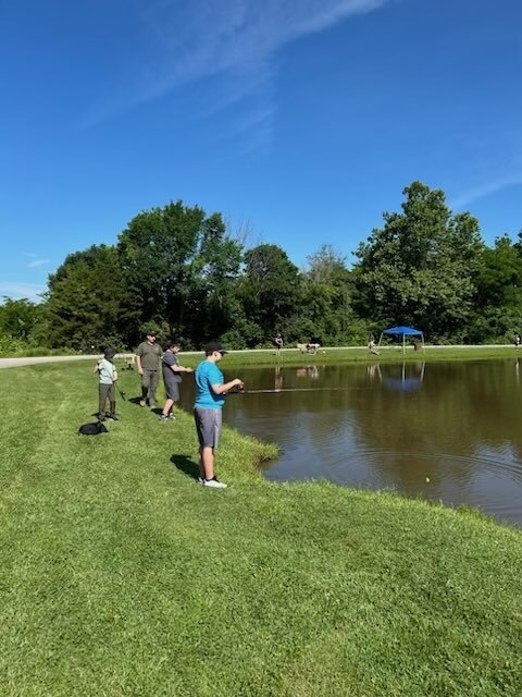 Students and instructors fish along the edge of a pond on a sunny day, practicing casting and fishing skills in a park setting.