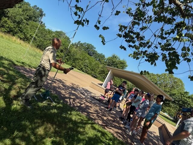 Students gather outdoors around a park ranger demonstrating fishing techniques during an educational field trip at a shaded park pavilion.