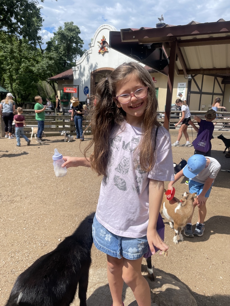 Student smiles while holding a bottle near a goat exhibit, with other children interacting with goats in the background.