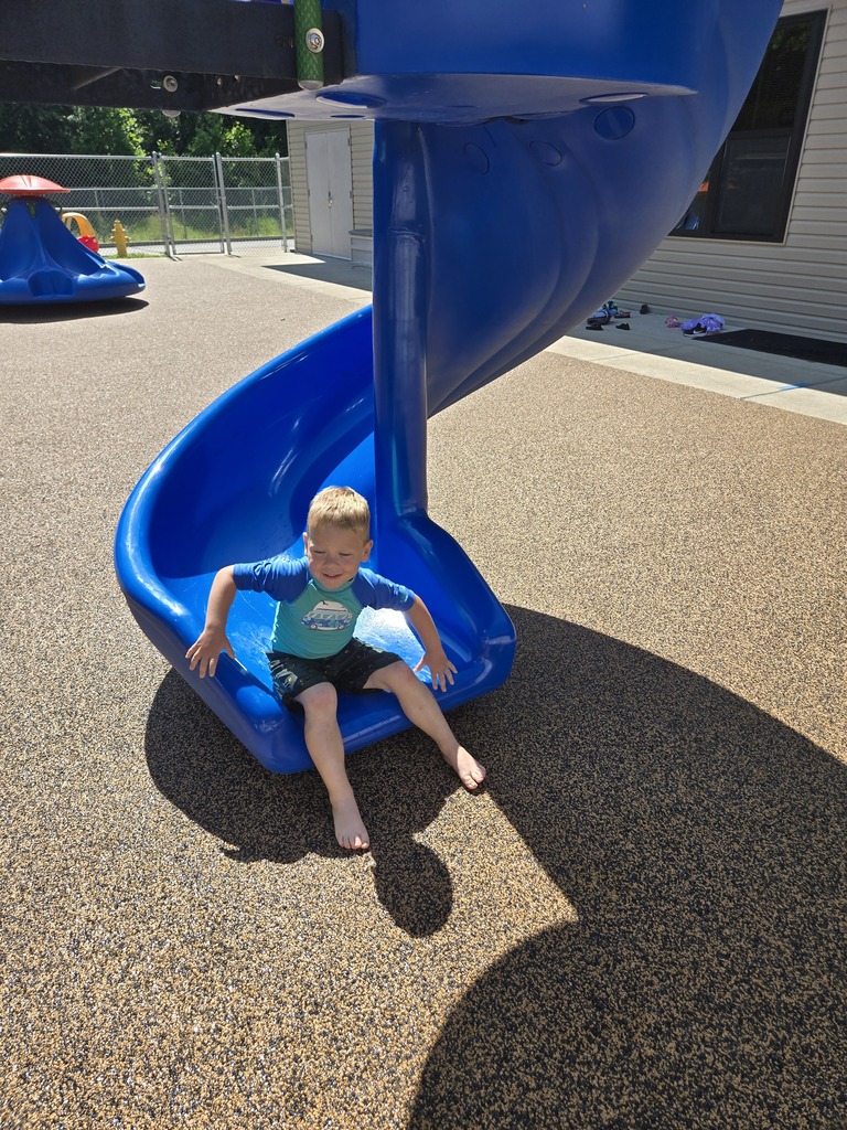 Young child smiles while sliding down a bright blue playground slide, barefoot and wet from water play.