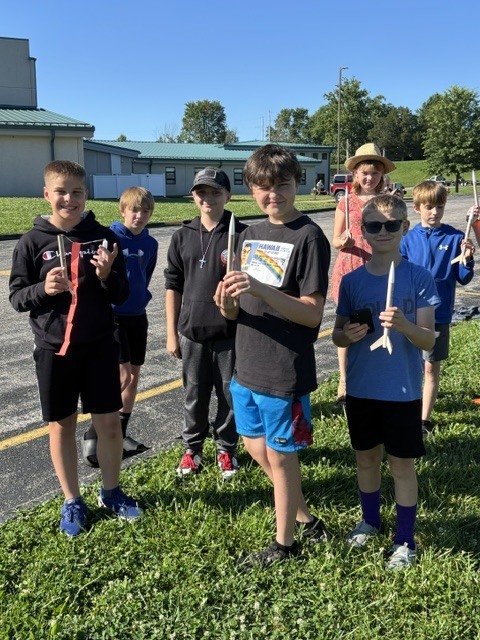 Students stand outdoors holding model rockets and launch equipment during a school rocket activity, smiling and preparing for a launch on a sunny day.