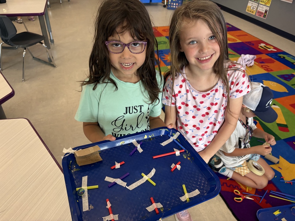 Two smiling students proudly display a tray featuring a handmade marble maze built with straws, tape, and cardboard during a classroom STEM project.