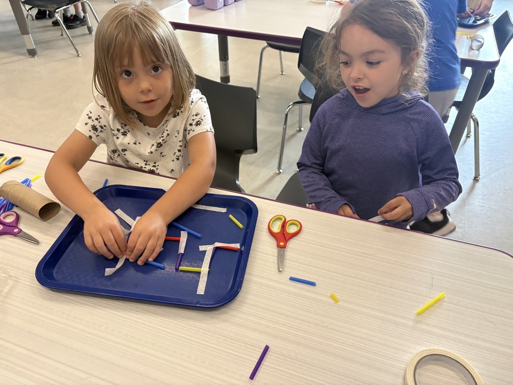 Two young students work together at a classroom table, arranging colorful straw pieces and tape on a tray during a hands-on STEM activity.