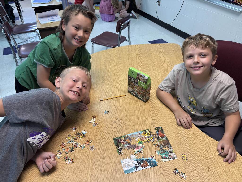 Three smiling students gather around a classroom table, working together to complete a colorful jigsaw puzzle.