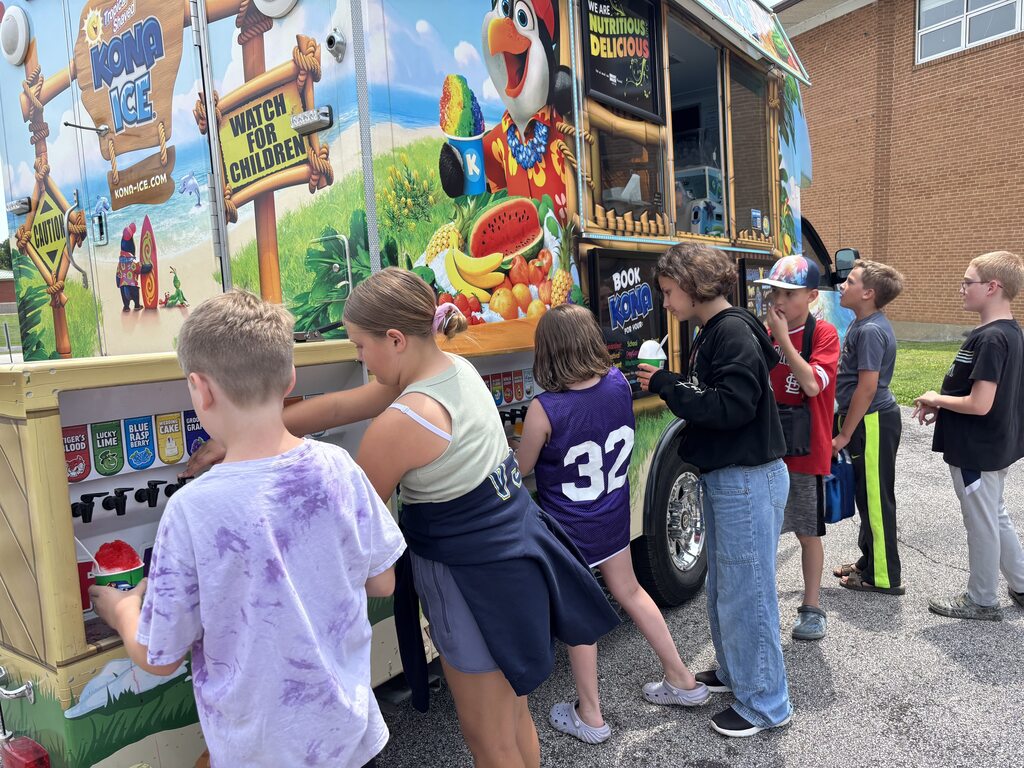 Students line up at a Kona Ice truck, choosing colorful shaved ice treats during an outdoor school event.
