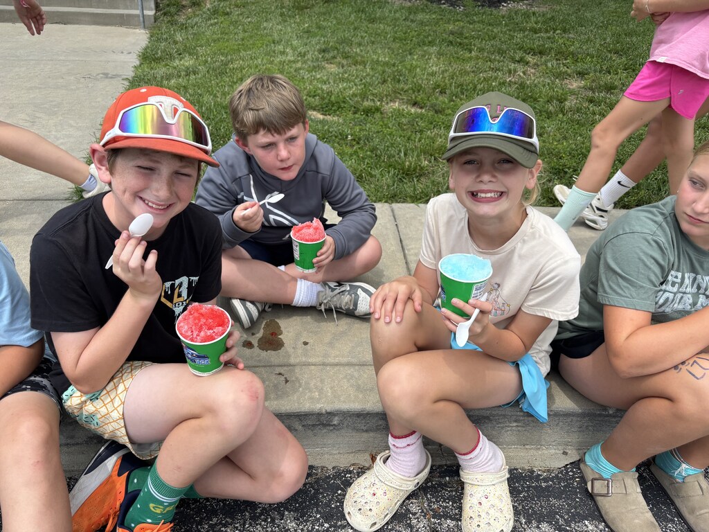 Students sit together on a curb, smiling and enjoying colorful shaved ice treats on a warm day.