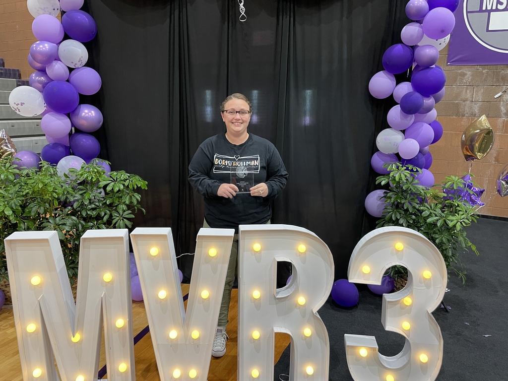 Staff member holds an award while standing between purple balloon arches behind illuminated MVR-3 letters at a recognition event.