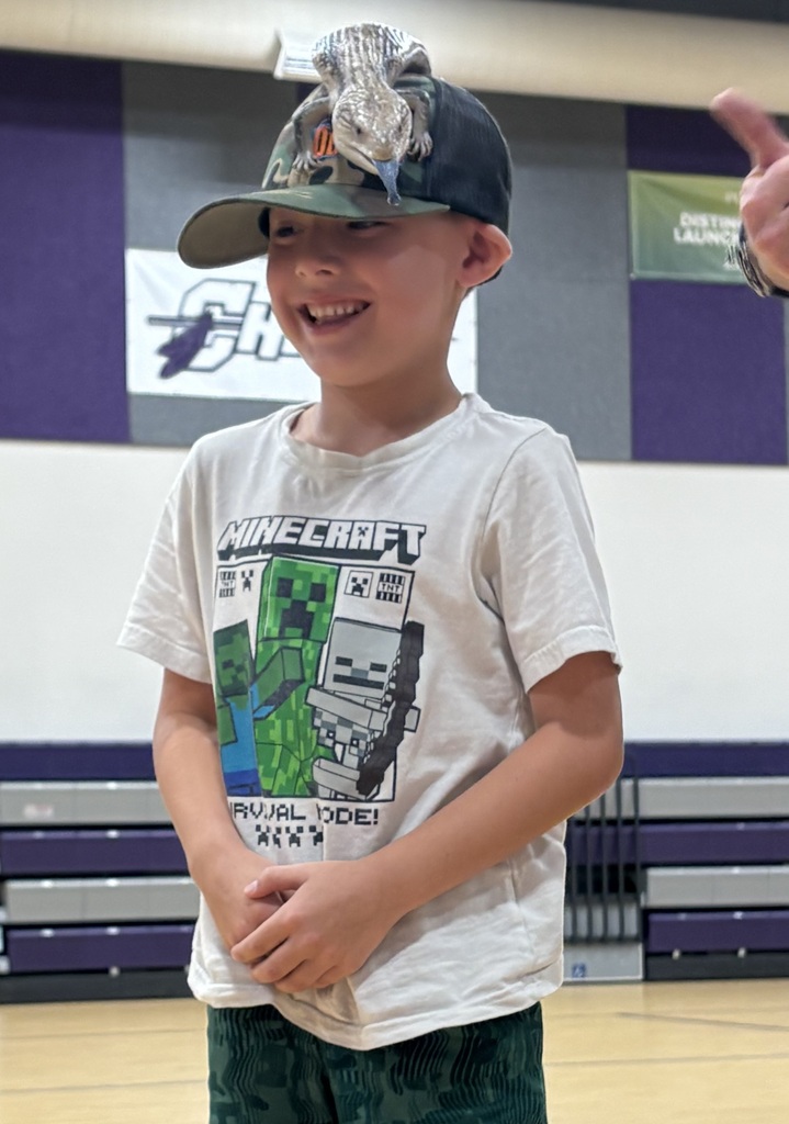 Smiling student stands in a school gym while a lizard rests on top of their baseball cap during a live animal presentation.