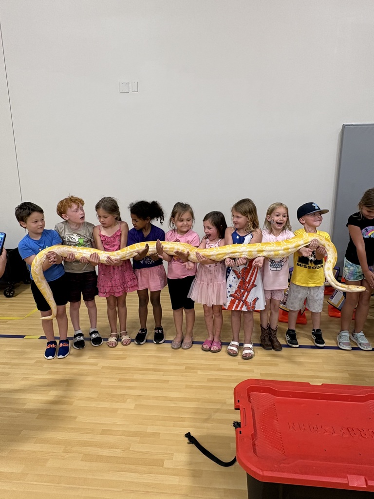A group of students stands in a line, smiling and helping hold a large albino python during an educational animal program.