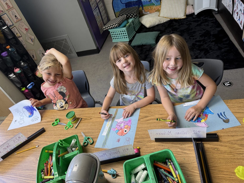 Three kindergarten students sit at a classroom table, smiling as they color fish drawings with crayons during an art activity.
