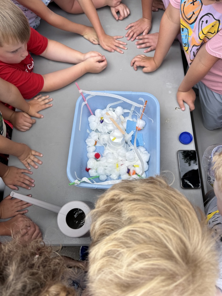 Children gather around a sensory bin filled with cotton balls, ribbons, pom-poms, and craft materials, exploring and observing together.