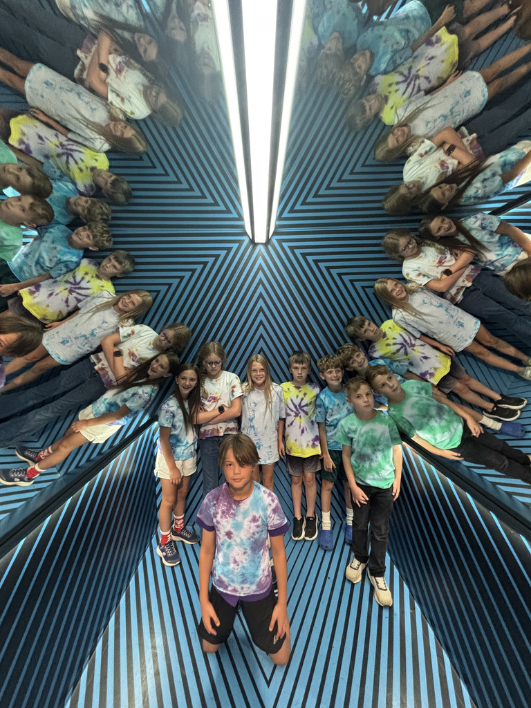 Students in tie-dye shirts pose inside a mirrored illusion room, creating repeating reflections along blue geometric walls.