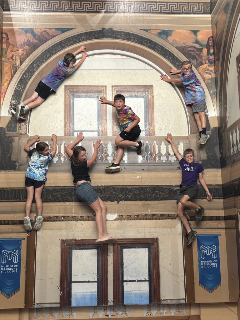 Students pose on an illusion exhibit wall, appearing to climb and hang from a grand building façade.