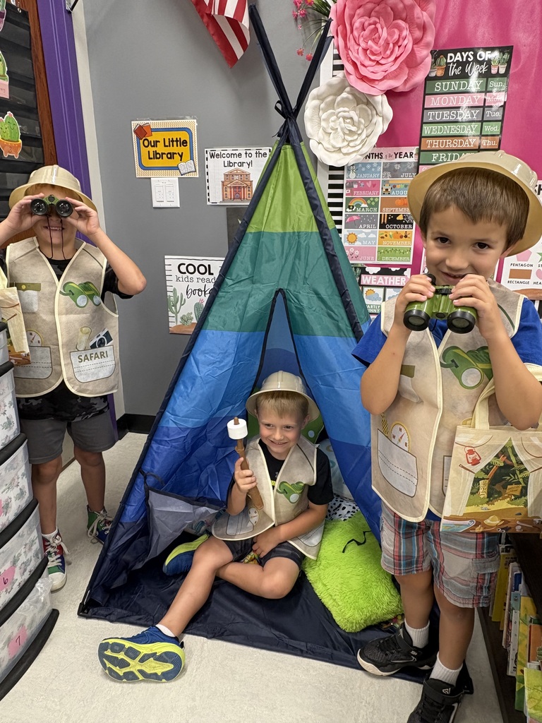 Three students dressed as safari explorers play in a classroom reading corner with a tent, binoculars, and camping props.