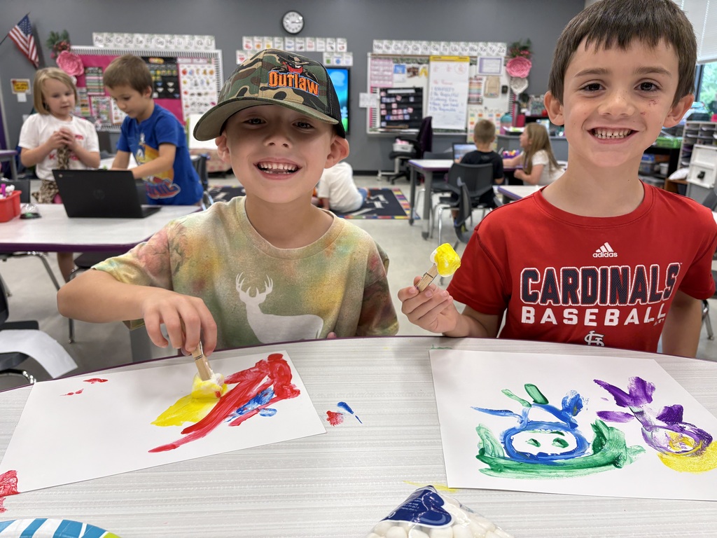 Two students smile while painting colorful artwork with paintbrushes during a classroom art activity.