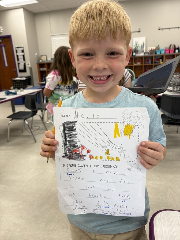 Student proudly holds up a camping-themed writing and drawing assignment while smiling in the classroom.