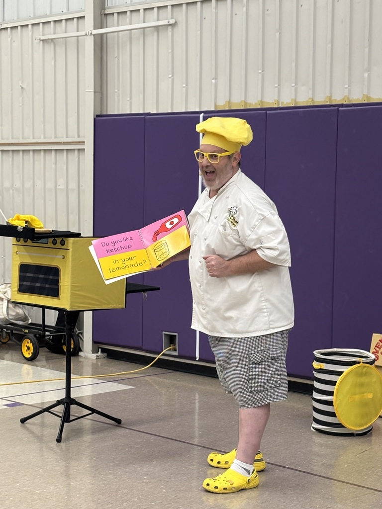 A chef-themed performer reads a humorous book aloud during a school assembly.