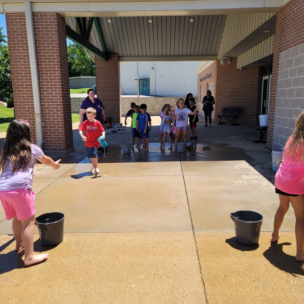Students participate in a water relay game outside, carrying and pouring water between buckets.