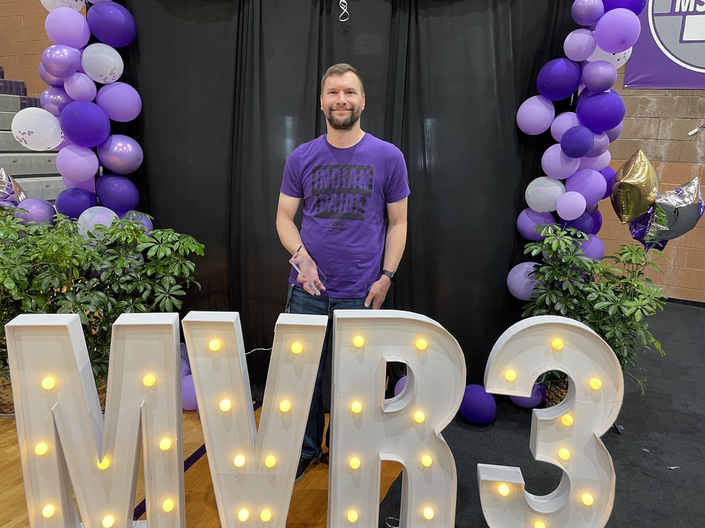 Person stands behind illuminated “MVR-3” letters at an awards event, framed by purple balloon arches and greenery.