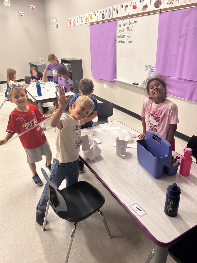 Students proudly pose beside a tall paper tower they built during a classroom STEM activity.