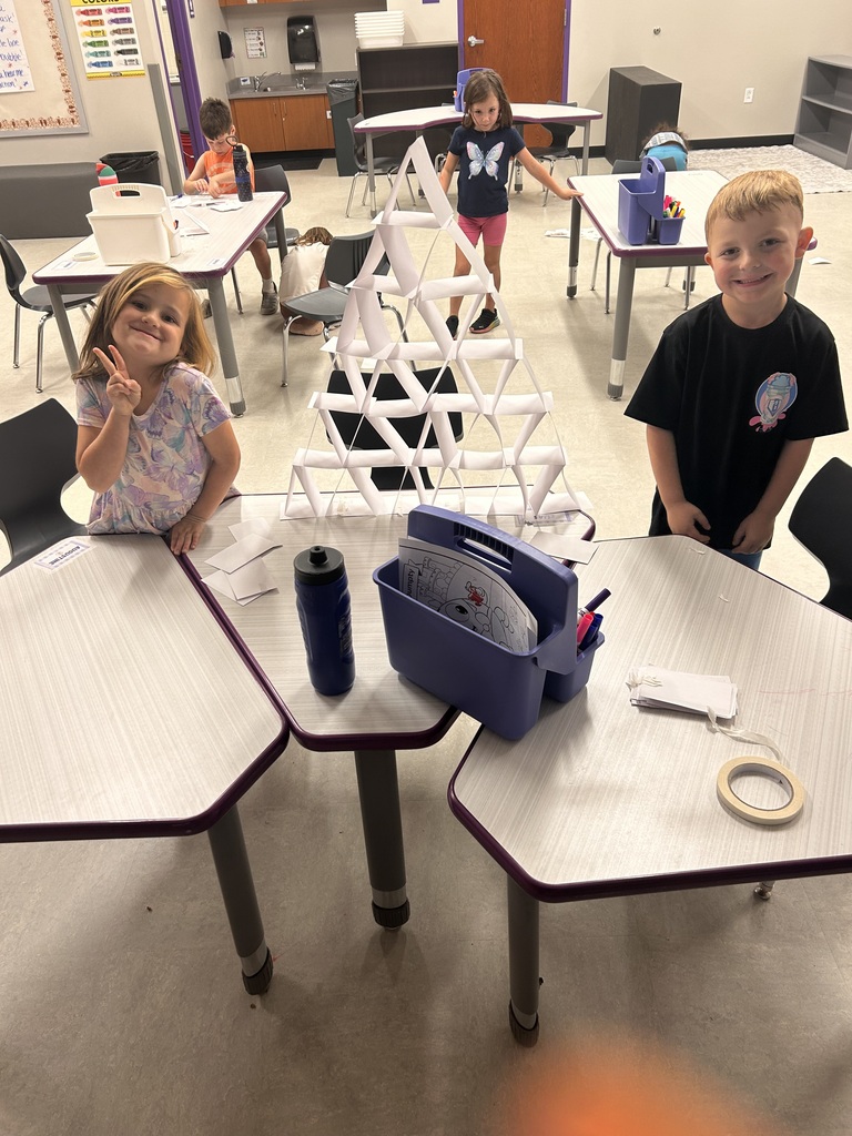 Students smile and wave while working together on a paper engineering challenge in an elementary classroom.