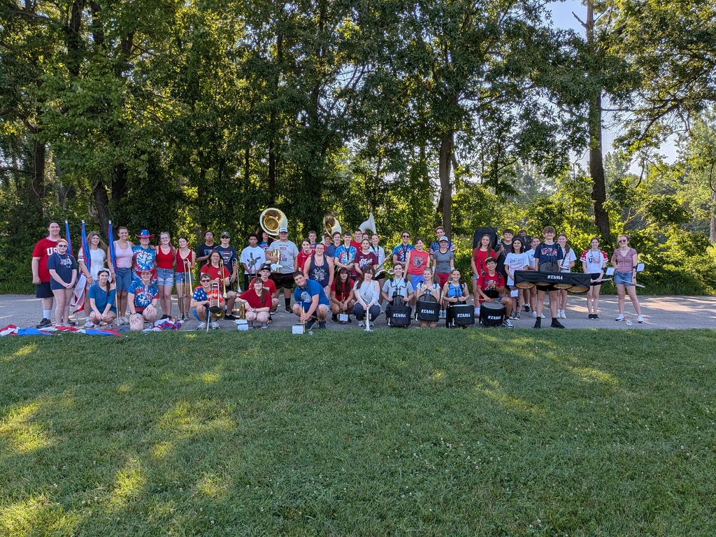 School marching band members pose outdoors with instruments and flags during summer practice on a sunny day.