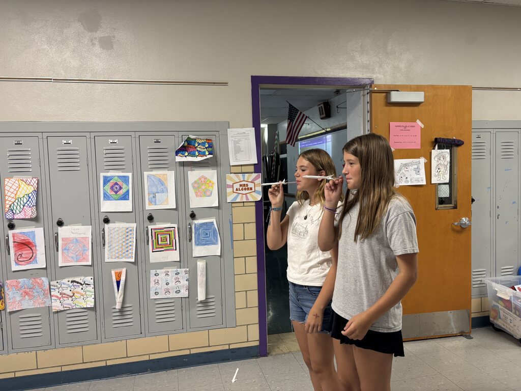 Two students prepare to launch paper airplanes during a classroom STEM activity, with student artwork displayed on nearby lockers.