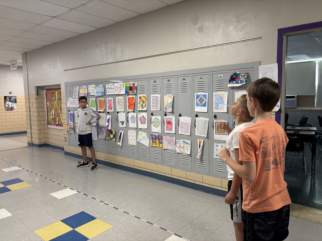 Students test handmade paper airplanes in a school hallway, launching them past lockers decorated with colorful geometric artwork.